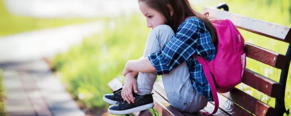 Przemoc domowa i przemoc psychiczna wobec dzieci – jak ją rozpoznać i przerwać? - smutna dziewczynka będąca ofiarą przemocy w szkole siedzie z plecakiem na ławce - blog Marita Woźny